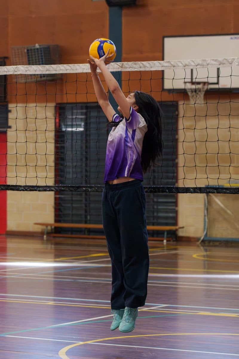 Girl in Obsidian jersey jumping to set a volleyball at the net in the Baulkham Hills hall
